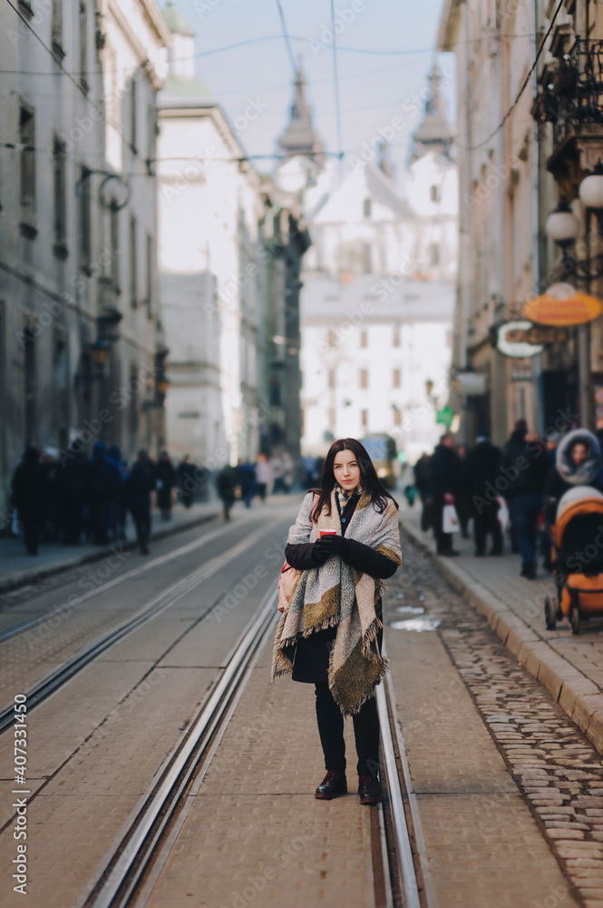 A young caucasian woman in an autumn coat stands in the middle of the streets on a tram line and drinks coffee surrounded by passers-by. Lviv, Ukraine.