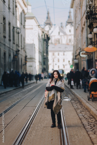 A young caucasian woman in an autumn coat stands in the middle of the streets on a tram line and drinks coffee surrounded by passers-by. Lviv, Ukraine. © shchus