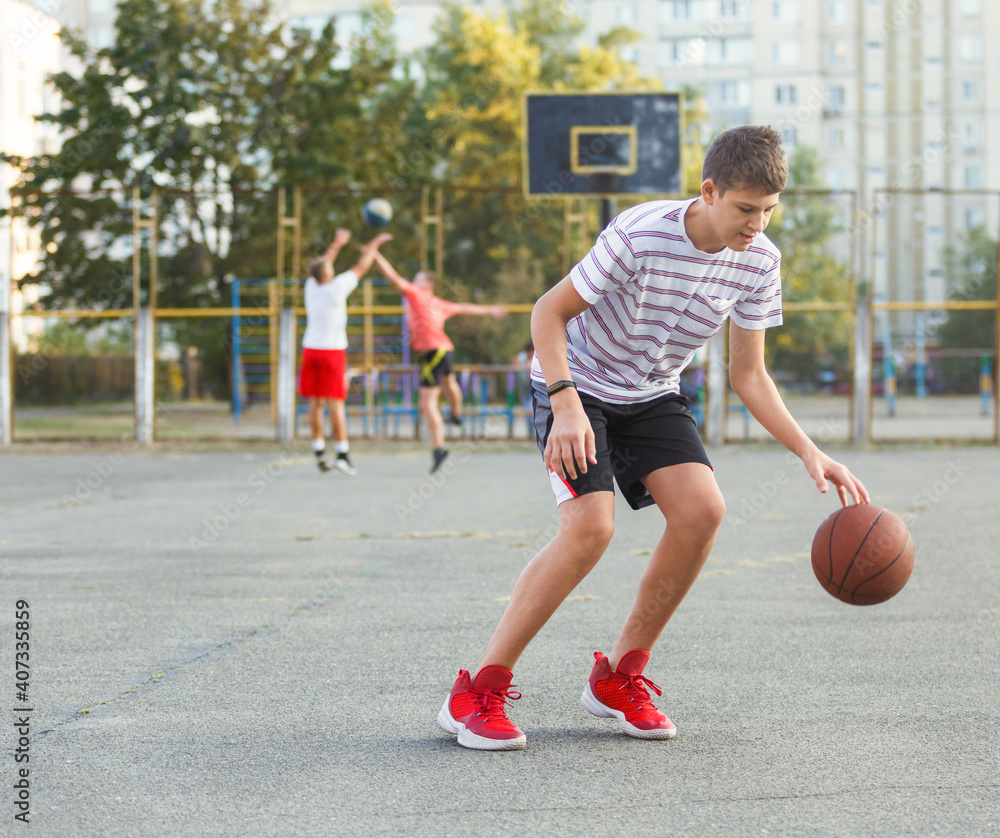 Young cute boy plays basketball at the outdoor streetball court on a sunny summer day. Teenager player in action dribbling the ball. Hobby, active lifestyle, sports for kids.