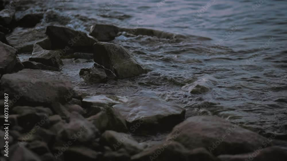 A static detail shot of small waves on the lake during dusk in slow motion  Summer evening on the riverside, stones washed by small waves.