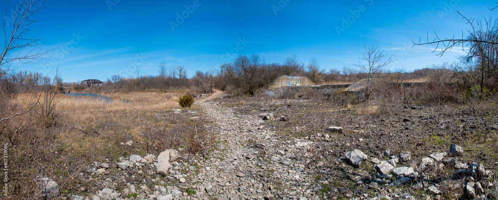 A rocky path leads through a deserted and overgrown field with an old bridge in the distance on a bright sunny day near Welland, Ontario.