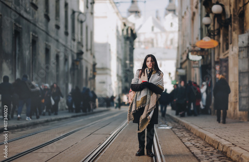A young caucasian girl in an autumn coat stands in the middle of the streets on a tram line and drinks coffee surrounded by passers-by. Lviv, Ukraine.