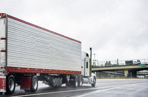 Industrial classic big rig white semi truck transporting cargo in refrigerator semi trailer running on the wide highway road with wet surface at raining weather