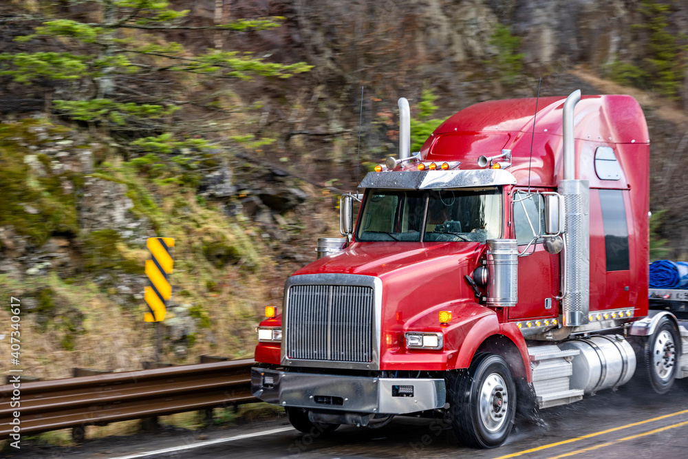 Industrial grade powerful red big rig semi truck with flat bed semi trailer running with rain dust on the wet road with protective fence on the side at raining weather