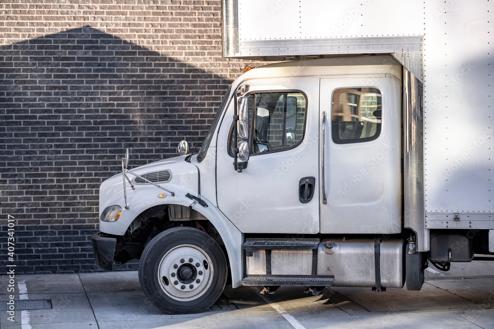 Medium power rig semi truck with long cabin and extended over the roof box trailer standing on the street with bricks apartment building Medium power rig semi truck with long cabin and extended over the roof box trailer standing on the street with bricks apartment building