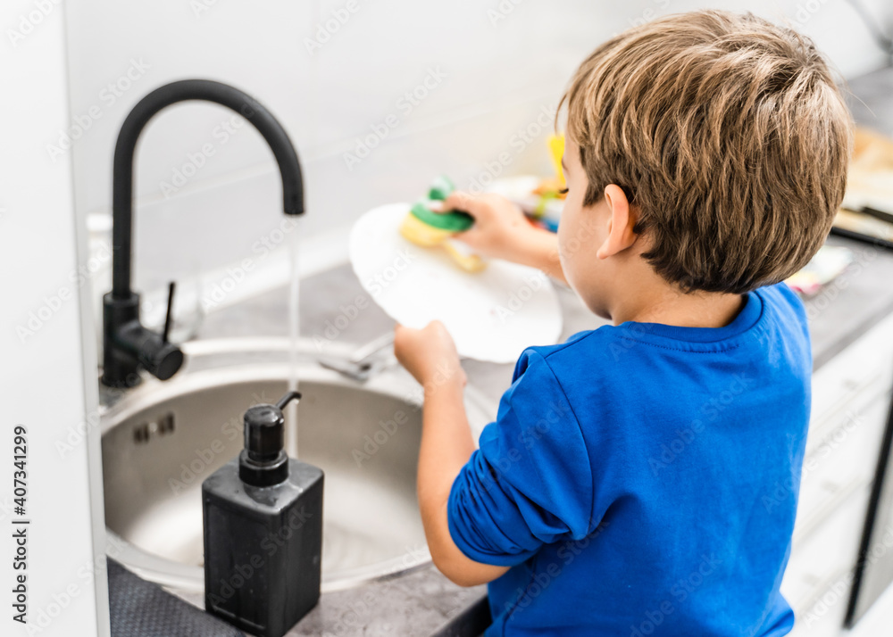 Back view of unknown caucasian boy or girl standing by the sink in the kitchen washing dishes - kid playing while helping in housework -growing up independence concept copy space Back view of unknown caucasian boy or girl standing by the sink in the kitchen washing dishes - kid playing while helping in housework -growing up independence concept copy space