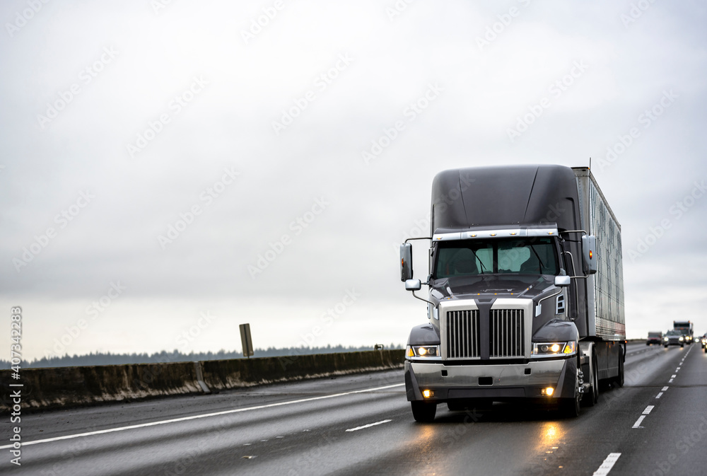 Stylish dark gray big rig semi truck with turned on headlights running with semi trailer on the straight evening wide highway road with light reflection