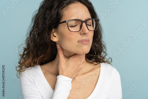 Woman having sore throat, tonsillitis, feeling sick, suffering from painful swallowing, angina, strong pain in throat, loss of voice, holding hand on her neck, isolated on studio blue background. photo