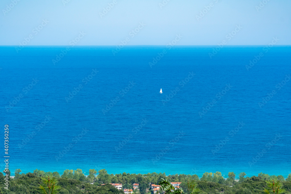 In far distance a single floating yacht with open white sails traveling in tranquil waters. Turquoise blue sea and sky background with copy space. Green trees and houses on bottom frame