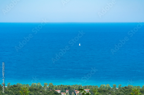 In far distance a single floating yacht with open white sails traveling in tranquil waters. Turquoise blue sea and sky background with copy space. Green trees and houses on bottom frame