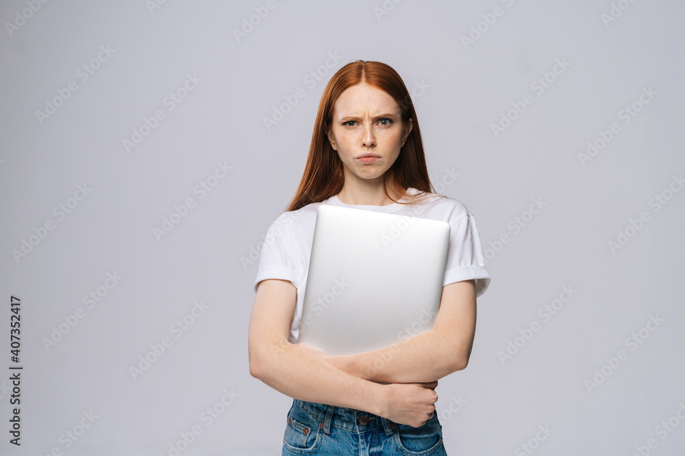 Distressed upset young business woman or student holding laptop computer and looking at camera on isolated gray background. Pretty lady model with red hair emotionally showing facial expressions. Distressed upset young business woman or student holding laptop computer and looking at camera on isolated gray background. Pretty lady model with red hair emotionally showing facial expressions.