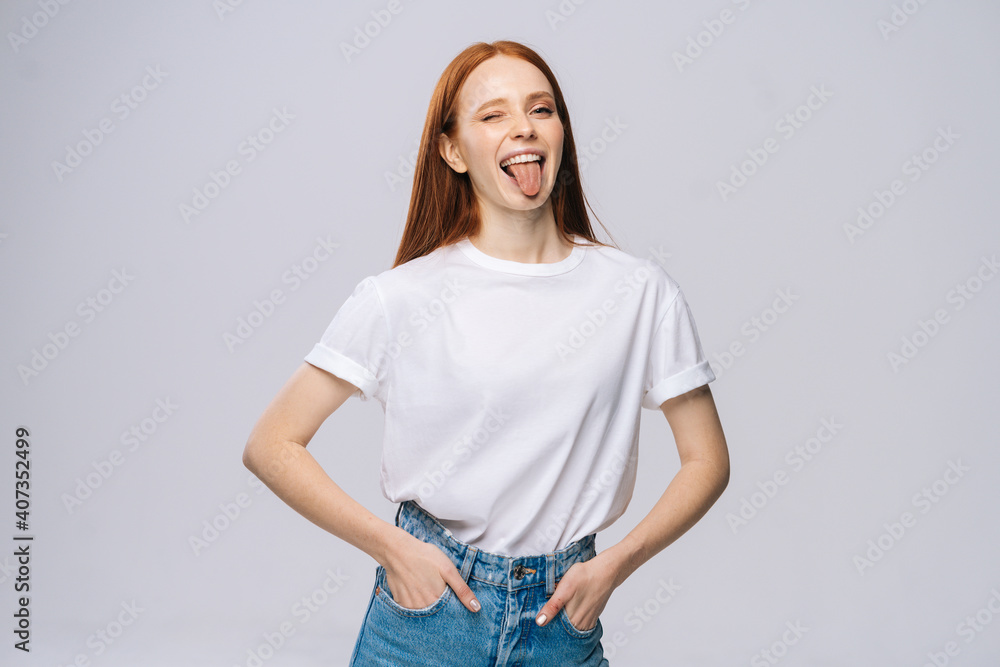 Obraz premium Joyful young woman wearing T-shirt and denim pants showing tongue on isolated white background. Pretty lady model with red hair emotionally showing facial expressions in studio, copyspace.