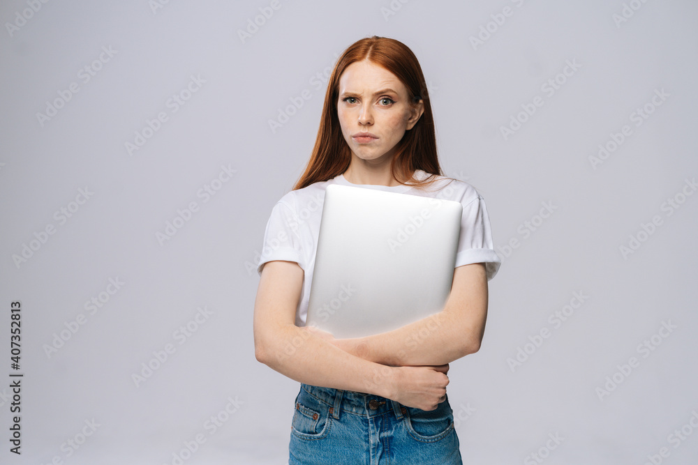 Frustrated upset young business woman or student holding laptop computer and looking at camera on isolated gray background. Pretty lady model with red hair emotionally showing facial expressions. Frustrated upset young business woman or student holding laptop computer and looking at camera on isolated gray background. Pretty lady model with red hair emotionally showing facial expressions.