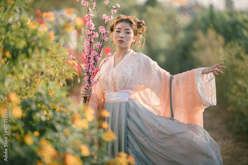 Portrait of beautiful asian young woman wearing  traditional cheongsam costume holding  cherry blossoms in garden. Costume chinese girl holding pink flower under green tree . photo