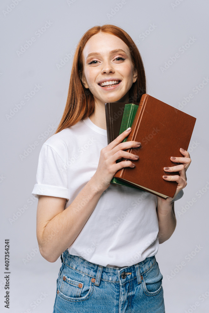 Cheerful young woman college student holding book and looking at camera on isolated gray background. Pretty redhead lady model wearing casual fashion clothes emotionally showing facial expressions. Cheerful young woman college student holding book and looking at camera on isolated gray background. Pretty redhead lady model wearing casual fashion clothes emotionally showing facial expressions.