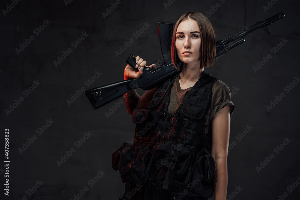 Dressed in black armour with dark shirt martial woman with short haircut poses in dark background holding assault rifle.