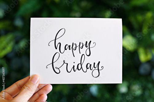 Hand holding card with modern calligraphy text "Happy Friday" on green nature background. 