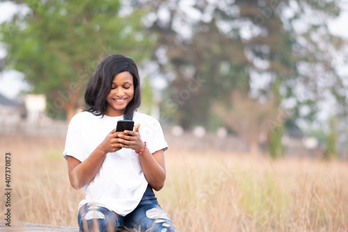 young and beautiful light skin african lady sitting a lone in a park smiling while using her phone