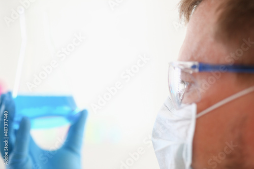 Fototapeta Naklejka Na Ścianę i Meble -  A portrait of a young surgeon chemist's doctor looks at a container with a blue liquid and a mask is fought with viruses and a vaccine for vaccines against diseases