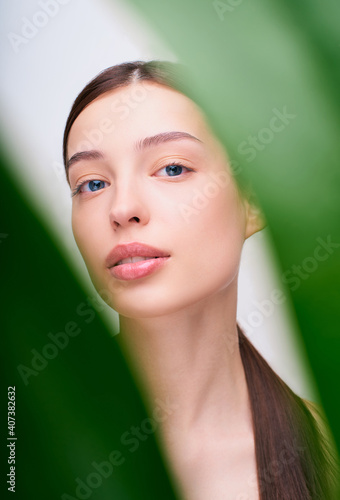 A cute woman's face with nude makeup peeps through the green leaves.