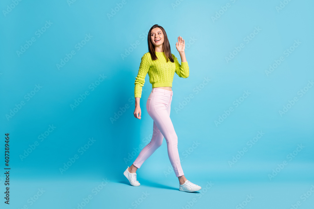 Full size profile side photo of young woman smile go walk wave hand hello hi look empty space isolated over blue color background Full size profile side photo of young woman smile go walk wave hand hello hi look empty space isolated over blue color background