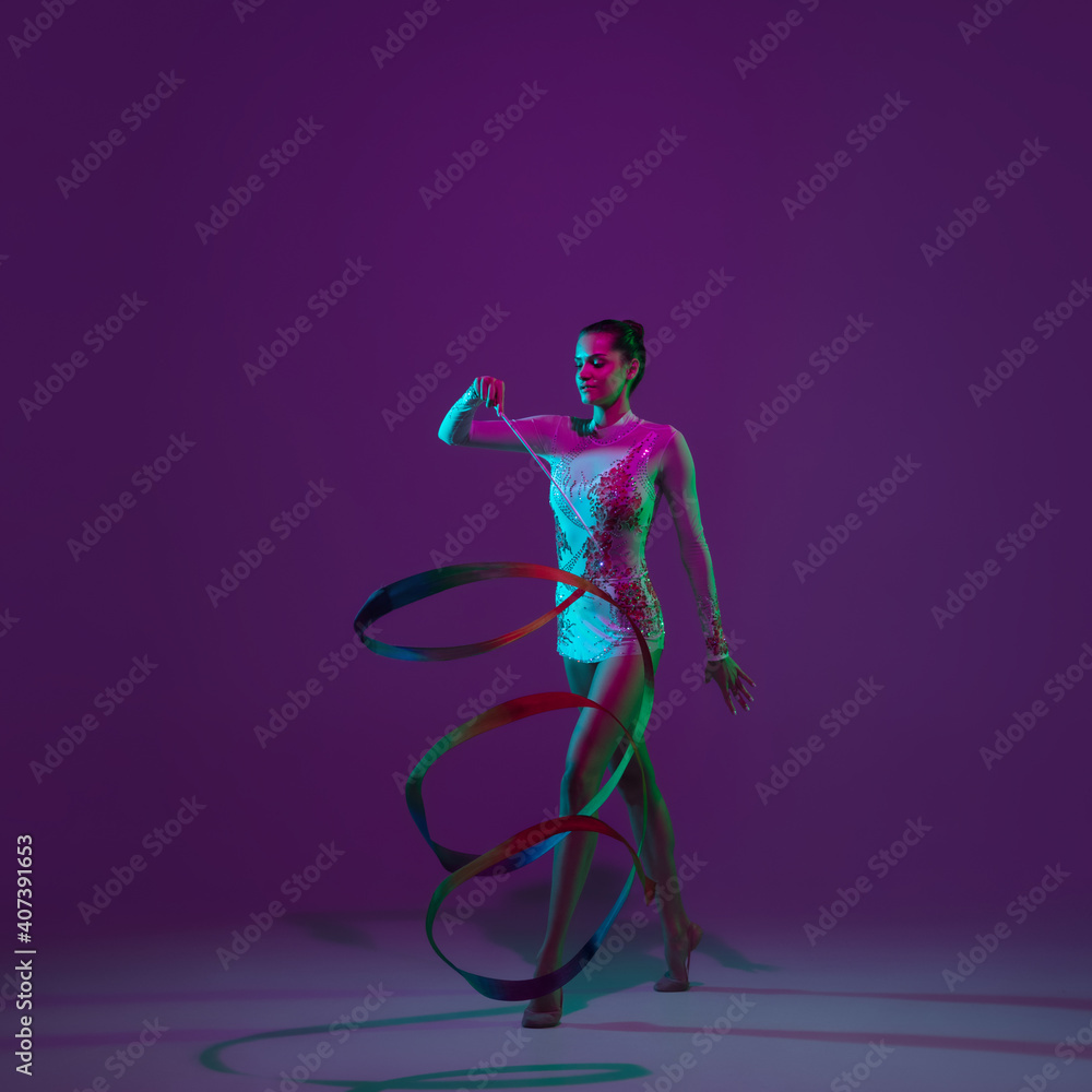 Motion. Young female athlete, rhythmic gymnastics artist dancing, training with tape isolated on purple studio background in neon light. Beautiful girl practicing with equipment. Grace in performance.