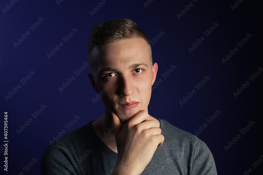 Young caucasian blonde man in grey t shirt on dark purple background touching his chin and thinking about something.