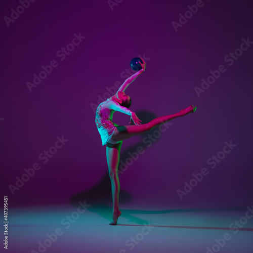 Free. Young female athlete, rhythmic gymnastics artist dancing, training with ball isolated on purple studio background in neon light. Beautiful girl practicing with equipment. Grace in performance.