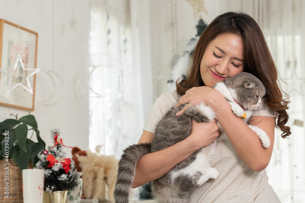 Happy Asian woman holding and hug her lovely fluffy grey and white cat in her arm at home. Cute kitten with yellow eyes and yellow bell collar celebration Christmas holiday with human