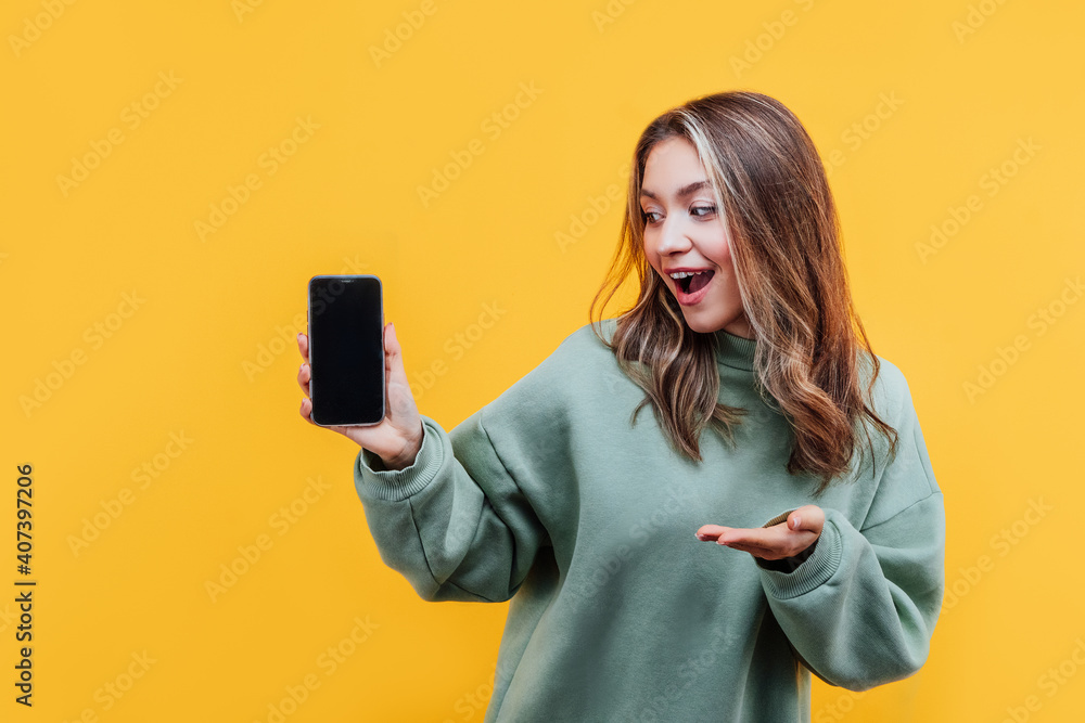 Studio photo of a girl on a yellow background. The girl holds a phone with a black screen and the other hand points to the screen Studio photo of a girl on a yellow background. The girl holds a phone with a black screen and the other hand points to the screen