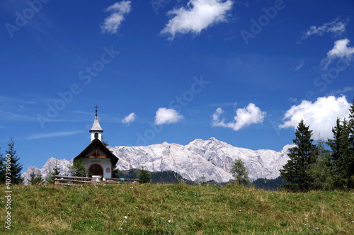 Kleine Bergkapelle auf einer Bergwiese mit gewaltigem Gebirgsmassiv im Hintergrund unter blauem Himmel