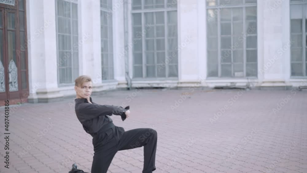 Wide shot of slim handsome ballet dancer spinning to camera and sitting down on one knee. Portrait of confident handsome young Caucasian man performing in urban city outdoors. Wide shot of slim handsome ballet dancer spinning to camera and sitting down on one knee. Portrait of confident handsome young Caucasian man performing in urban city outdoors.