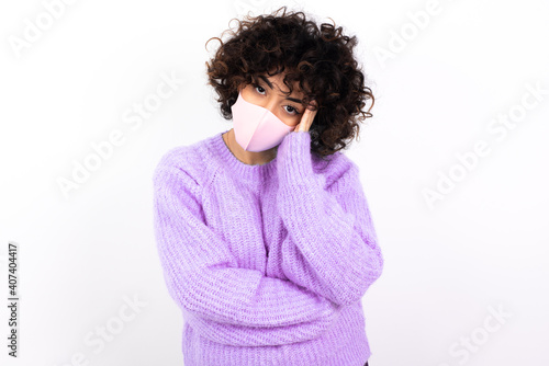Very bored young beautiful caucasian woman wearing medical mask standing against white wall holding hand on cheek while support it with another crossed hand, looking tired and sick, © Roquillo