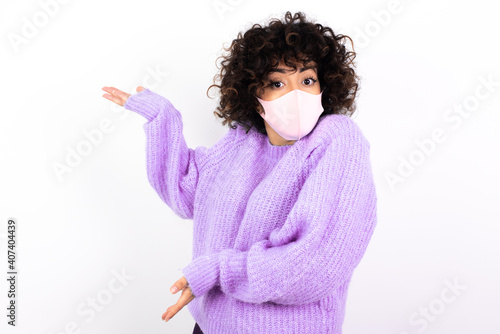 young beautiful caucasian woman wearing medical mask standing against white wall pointing aside with both hands showing something strange and saying: I don't know what is this. Advertisement concept. © Roquillo