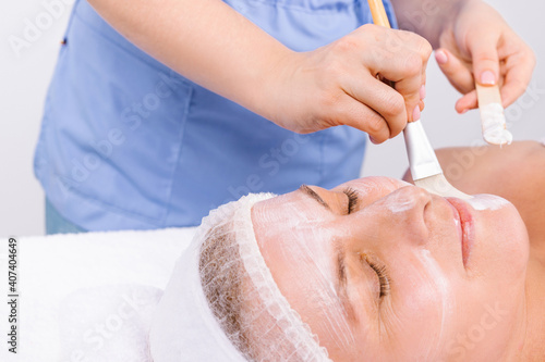 Cropped photo of a beautician applying a moisturizing face mask with a cosmetic brush to a senior woman a headband bandage on her head . Spa and beauty concept