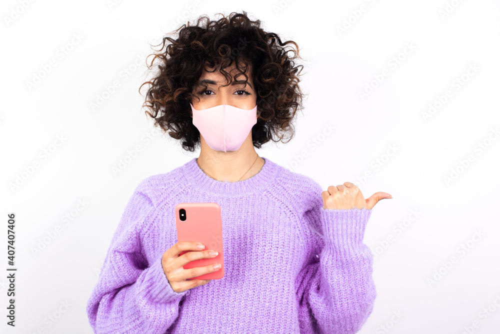young beautiful caucasian woman wearing medical mask standing against white wall using and texting with smartphone pointing and showing with thumb up to the side with happy face smiling young beautiful caucasian woman wearing medical mask standing against white wall using and texting with smartphone pointing and showing with thumb up to the side with happy face smiling