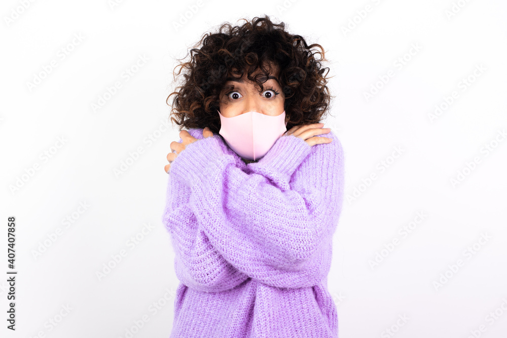 Desperate young beautiful caucasian woman wearing medical mask standing against white wall trembles and feels cold, hugs oneself to warm up or feels scared notices something terrifying. Desperate young beautiful caucasian woman wearing medical mask standing against white wall trembles and feels cold, hugs oneself to warm up or feels scared notices something terrifying.