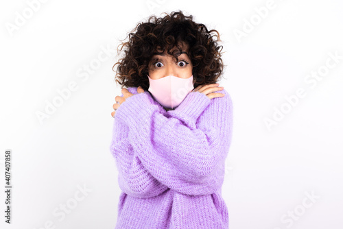 Desperate young beautiful caucasian woman wearing medical mask standing against white wall trembles and feels cold, hugs oneself to warm up or feels scared notices something terrifying. © Roquillo