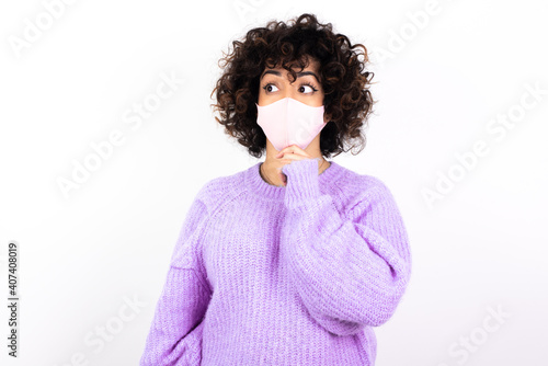 Thoughtful young beautiful caucasian woman wearing medical mask standing against white wall holds chin and looks away pensively makes up great plan © Roquillo