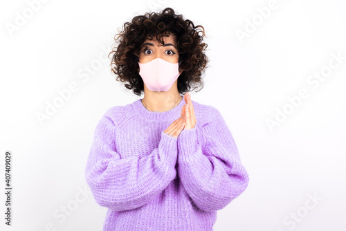 Surprised emotional young beautiful caucasian woman wearing medical mask standing against white wall rubs palms and stares at camera with disbelief © Roquillo