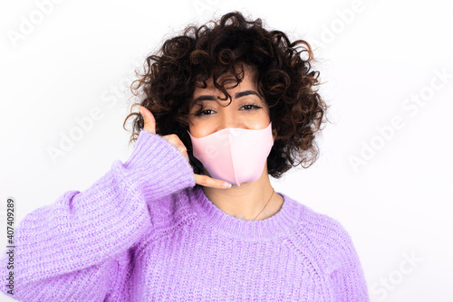 young beautiful caucasian woman wearing medical mask standing against white wall makes phone gesture, says call me back again, has glad expression.