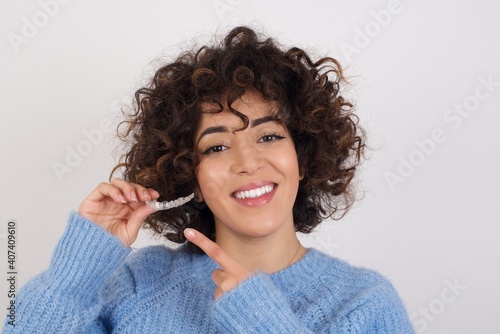young beautiful caucasian woman wearing blue knitted sweater standing against white studio background holding an invisible aligner braces and smiling. Dental healthcare concept. 