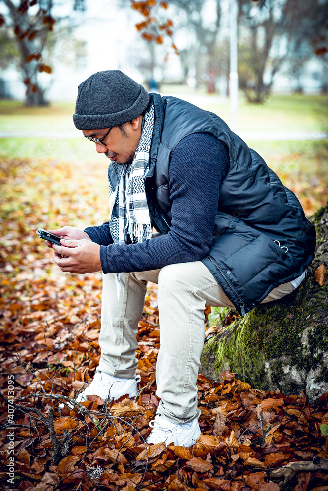 Asian man sitting outside under a big tree with his mobile phone Asian man sitting outside under a big tree with his mobile phone