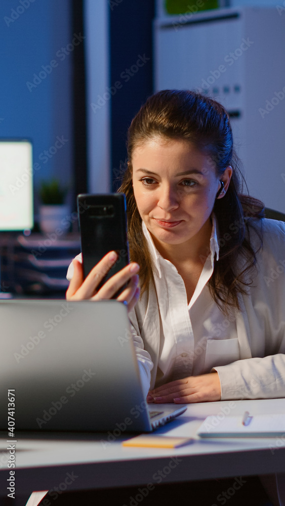 Business woman discussing during video conference with partners using smartphone and headphone late at night working in start-up office. Employee with modern technology network wireless doing overtime Business woman discussing during video conference with partners using smartphone and headphone late at night working in start-up office. Employee with modern technology network wireless doing overtime