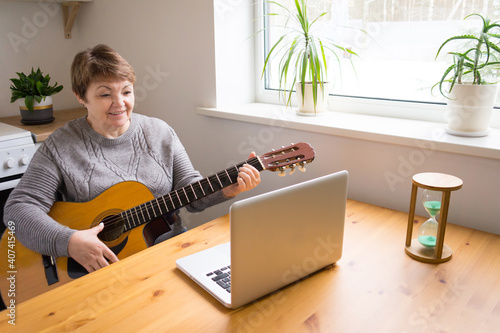 An elderly woman takes guitar lessons online. A retired senior woman studying online, watching music lessons at home on a laptop. Distance education