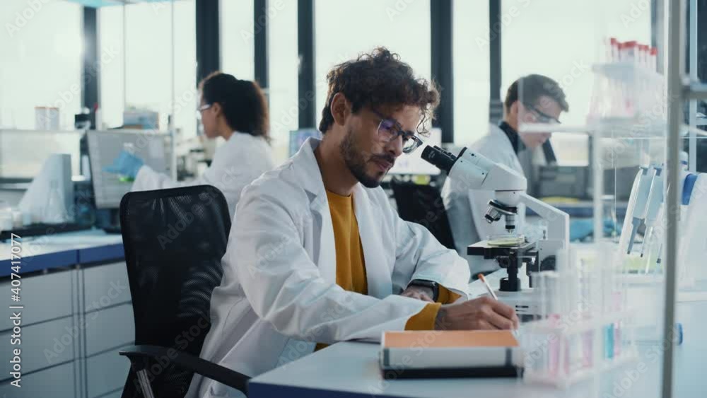 Medical Science Laboratory: Handsome Hispanic Scientist Looking Under Microscope Does Analysis of Test Sample. Diverse Team of Young Specialists, Using Advanced Technology Equipment. Side View Shot Medical Science Laboratory: Handsome Hispanic Scientist Looking Under Microscope Does Analysis of Test Sample. Diverse Team of Young Specialists, Using Advanced Technology Equipment. Side View Shot