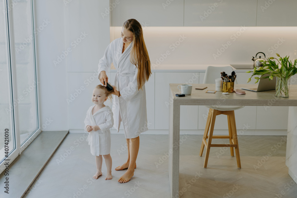 Caring mother combs hair of her small daughter wear white bathrobes pose against kitchen interior stand bare feet at floor. Beautiful little curly girl after taking shower. Mom making hairstyle to kid Caring mother combs hair of her small daughter wear white bathrobes pose against kitchen interior stand bare feet at floor. Beautiful little curly girl after taking shower. Mom making hairstyle to kid