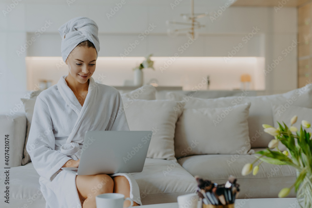 Horizontal shot of pretty young woman dressed in bathrobe and towel after shower sits on comfortabe sofa in living room works on laptop computer has own beauty blog types feedback to followers Horizontal shot of pretty young woman dressed in bathrobe and towel after shower sits on comfortabe sofa in living room works on laptop computer has own beauty blog types feedback to followers