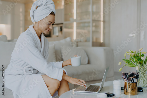 Indoor shot of female freelancer workds distantly rom home types on laptop computer dressed in bathrobe drinks coffee or tea enjoys domestic atmosphere surfs internet reads advice about skin care