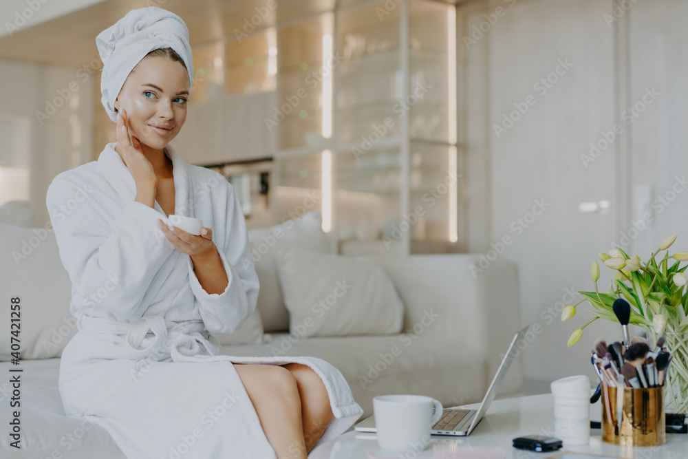 Horizontal shot of pretty Caucasian woman applies facial cream looks thoughtfully aside holds bar of cosmetic product dressed in bathrobe and towel sits on comfortable sofa over home interior Horizontal shot of pretty Caucasian woman applies facial cream looks thoughtfully aside holds bar of cosmetic product dressed in bathrobe and towel sits on comfortable sofa over home interior